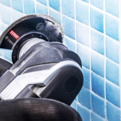 Esmerilhadora elétrica a trabalhar numa parede de azulejos azuis, mãos com luvas pretas a segurar a ferramenta.