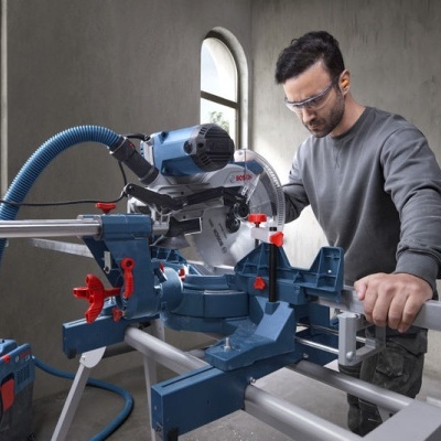 Serra de esquadria azul com detalhes vermelhos e pretos numa oficina com homem a usar óculos de proteção