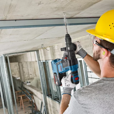 Homem com capacete amarelo a furar teto de concreto com furadeira azul e preta em canteiro de obras