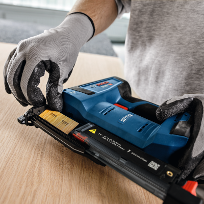 Pistola de pregos azul e preta em uso sobre madeira clara com luvas