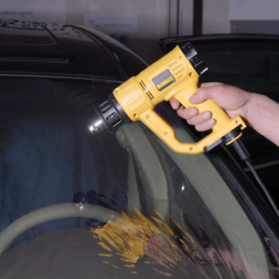 Pistola de ar quente amarela usada num vidro de carro preto em garagem