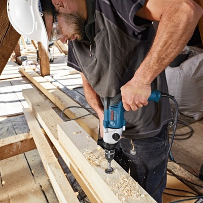 Homem a usar berbequim elétrico azul para perfurar madeira em sótão com vigas