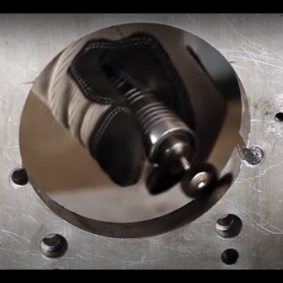 Ferramenta metálica redonda sobre base metálica com parafusos, segurada por luva preta.