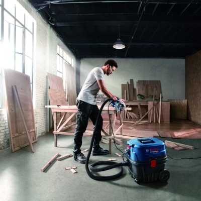 Aspirador industrial Bosch azul e preto numa oficina de carpintaria com tábuas de madeira e uma pessoa a usá-lo