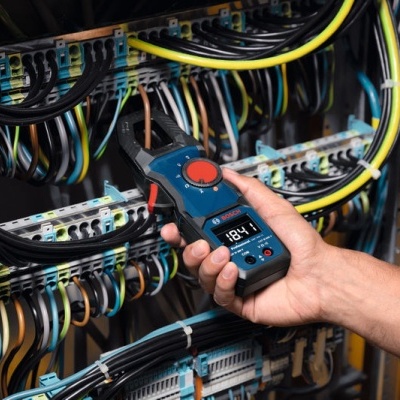 Multímetro digital azul e preto a medir voltagem em painel elétrico com muitos cabos