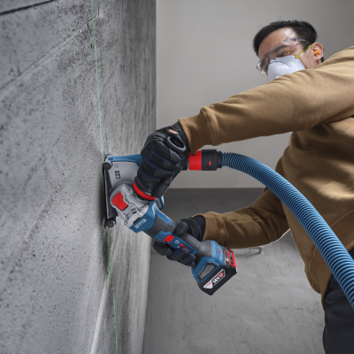 Pessoa a cortar parede cinzenta com cortadora elétrica Bosch azul e vermelha com tubo de aspiração azul.