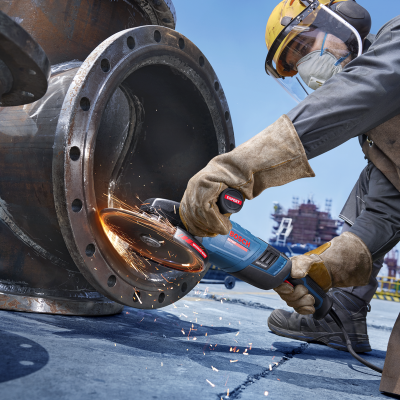 Trabalhador usa ferramenta angular Bosch azul para desbastar metal, cria faíscas, com luvas, capacete amarelo, máscara e viseira em ambiente industrial.