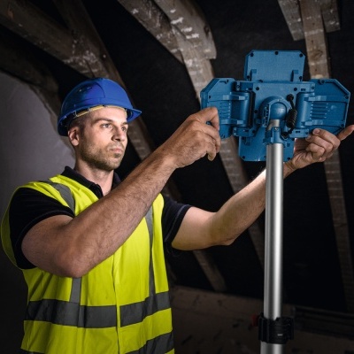 Homem com capacete azul e colete de segurança amarelo ajusta dispositivo azul num tripé de metal