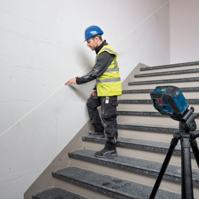Homem a medir parede numa escadaria com colete reflexivo e capacete azul, equipamento laser em tripé