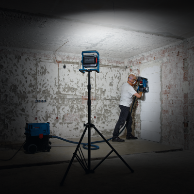 Lampião de obra azul em tripé e homem usando máquina elétrica azul para perfurar parede