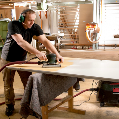 Homem a lixar placa branca numa oficina de carpintaria com máquinas ao fundo