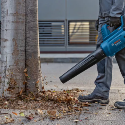 Soprador de folhas azul com detalhes pretos ao lado de árvore e folhas secas na calçada