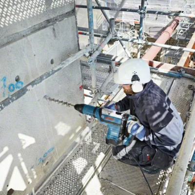 Operário com capacete e luvas perfurando parede cinza em andaime metálico