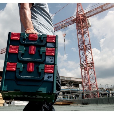 Caixa de ferramentas modular verde escura com fechos vermelhos numa obra com grua e edifícios