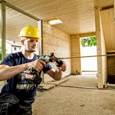 Homem com capacete amarelo a usar furadeira num interior de construção em madeira.