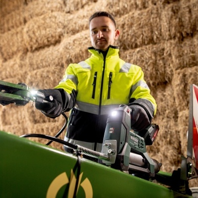 Homem com casaco amarelo de alta visibilidade e luvas operando máquina verde dentro de celeiro com fardos de palha.