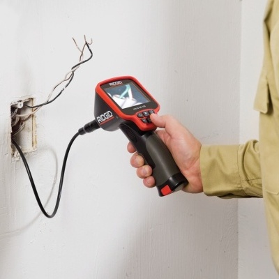Mão segura câmara de inspeção RIDGID com ecrã digital perto de caixa elétrica na parede