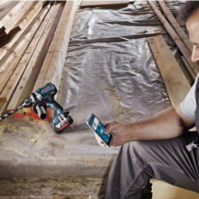 Homem segurando smartphone ao lado de berbequim elétrico em ambiente de construção de madeira.