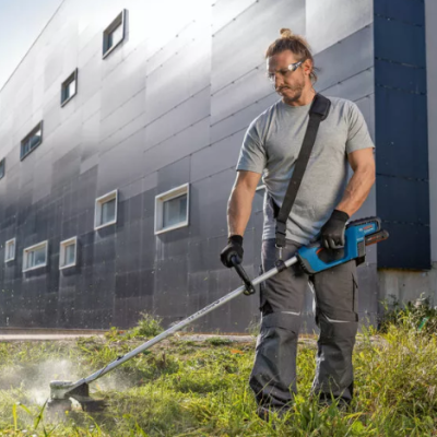 Homem a aparar relva com aparador sem fios azul e preto num espaço exterior com edifício cinzento