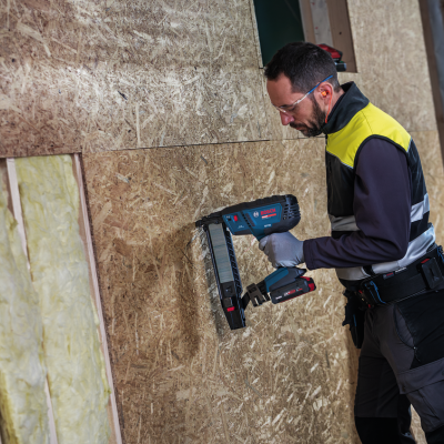 Homem a usar pistola de pregos Bosch em painéis de madeira OSB num ambiente de construção com isolamento.