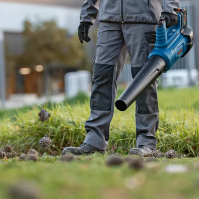 Pessoa com soprador de folhas azul e preto num jardim com relva e pinhas