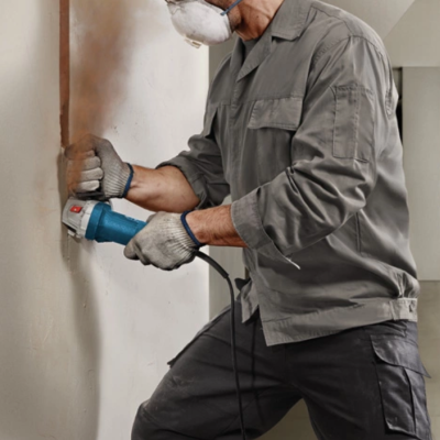 Homem a lixar parede com lixadora eletrica azul com roupa de trabalho cinza e máscara
