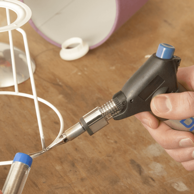 Soldador a gás portátil Dremel preto e azul em uso numa bancada de madeira com suporte branco e panela roxa ao fundo.