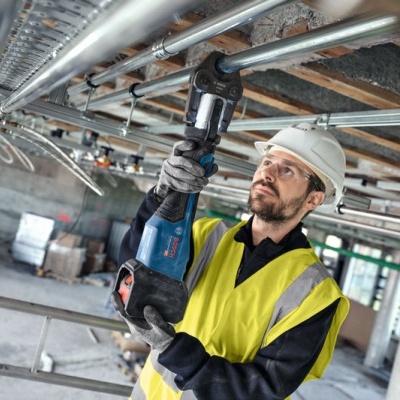 Homem com colete amarelo e capacete usa ferramenta elétrica azul para prender tubos no teto de construção