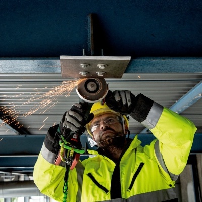 Homem a usar rebarbadora em estrutura metálica azul, vestido com colete amarelo e capacete.