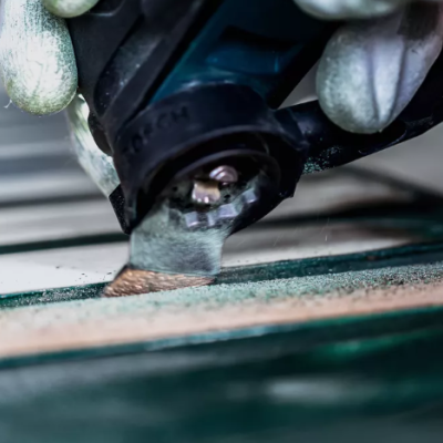 Mãos com luvas segurando ferramenta elétrica com lâmina cortando material verde e bege