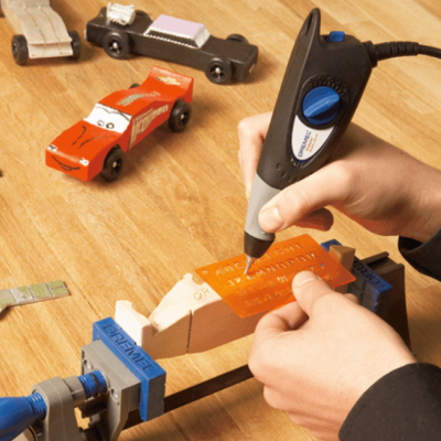 Mãos usam ferramenta de gravação numa peça de madeira laranja presa por morsa azul numa mesa de madeira com brinquedos e materiais de artesanato