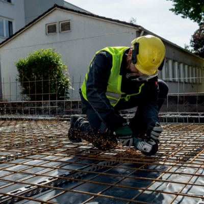 Trabalhador da construção civil com colete e capacete amarelo a manusear ferramenta elétrica sobre barras de metal