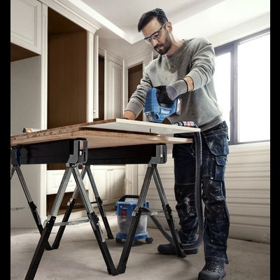 Homem a usar serra elétrica azul numa mesa de trabalho preta com madeira num quarto com chão cinzento e paredes brancas