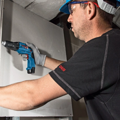 Homem com capacete azul usando furadeira elétrica para fixar placa de gesso