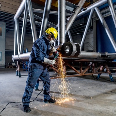 Trabalhador com capacete amarelo cortando peça metálica com faíscas em ambiente industrial.
