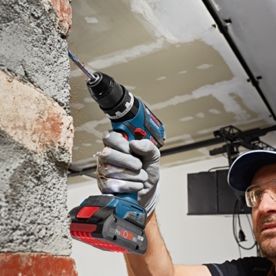 Homem usando furadeira elétrica azul trabalhando numa parede de tijolos e cimento