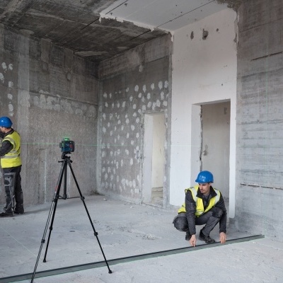 dois trabalhadores de construção civil com capacetes azuis e coletes refletivos medindo esquadro metálico em parede de cimento