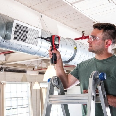 Pessoa a medir um tubo metálico de ventilação usando um dispositivo digital numa escada