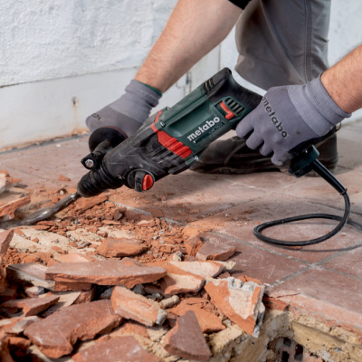 Martelo demolidor elétrico Metabo a quebrar azulejos castanhos no chão com parede branca ao fundo