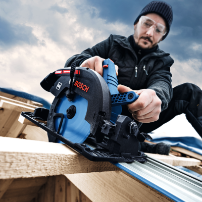 Serrote circular elétrico BOSCH com disco azul e detalhes a preto e vermelho, operado por homem com óculos de proteção, gorro e roupa escura, madeira clara e céu nublado.