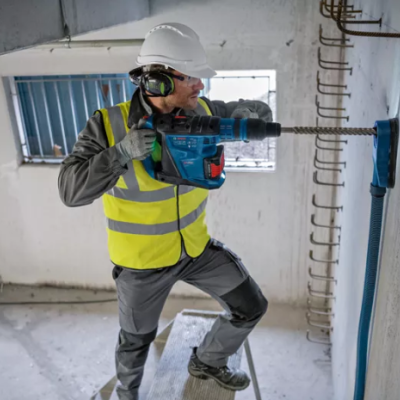 Homem com capacete e colete refletor a usar furadeira elétrica junto à parede num local de construção