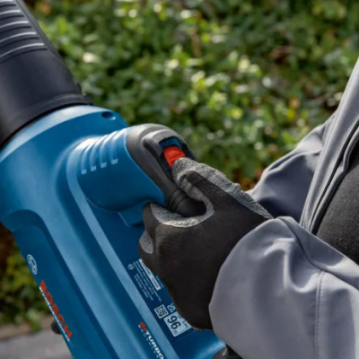 Mãos com luvas pretas seguram ferramenta elétrica azul com botão vermelho e fundo de plantas verdes