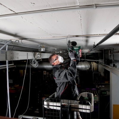 Homem com máscara e óculos a trabalhar com ferramenta elétrica num tecto baixo e estrutura metálica suspensa