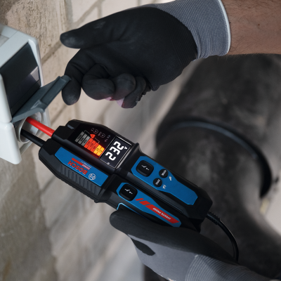 Tester de tensão Bosch com visor digital a mostrar 232 numa tomada branca numa parede de tijolo.