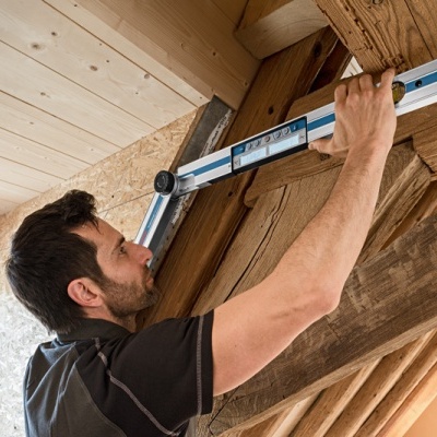 Homem mede estrutura de madeira rústica com nível de bolha azul num sótão de madeira clara