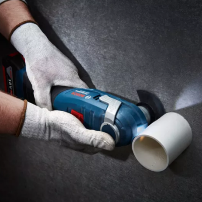 Ferramenta elétrica azul cortando tubo branco na parede cinza com luvas brancas