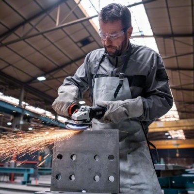 Homem a usar rebarbadora elétrica num bloco metálico dentro de oficina industrial.