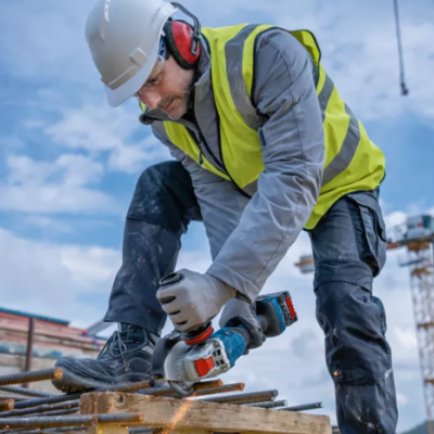 Trabalhador com capacete e colete a cortar barra de metal num canteiro de obras