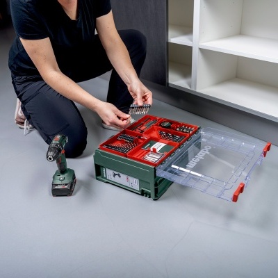 Organizador de ferramentas verde e vermelho com ferramentas em chão cinzento junto a móvel branco e cinza