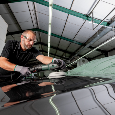 Homem polindo capot preto de carro com polidora elétrica numa oficina